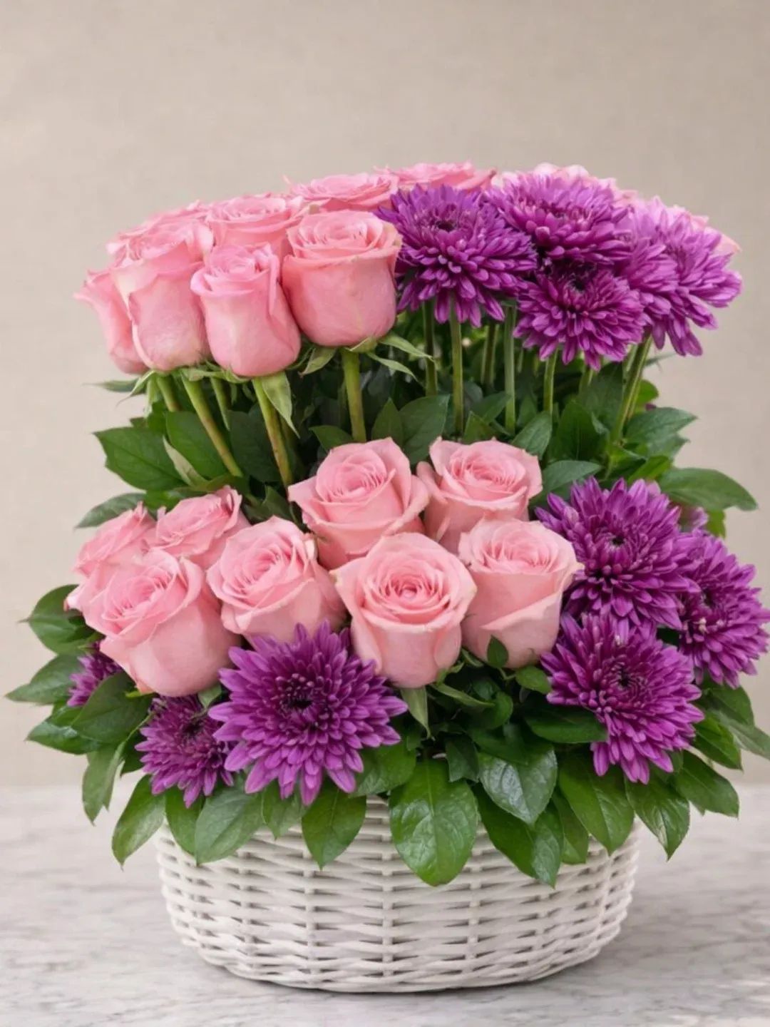 Pink roses and purple chrysanthemums arranged in a white wicker basket with green leaves